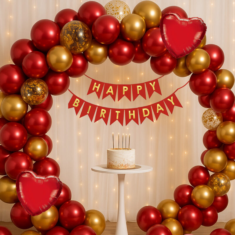 A celebratory setup featuring a Red and Gold Birthday Decor Kit, with a balloon arch made of red and gold metallic balloons, interspersed with gold confetti and red heart foil balloons, against a white net curtain backdrop.