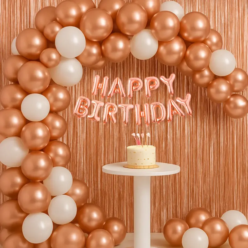 A beautifully decorated party setup using a Rose Gold Birthday Decor Kit, featuring a balloon arch made of rose gold and white balloons, a "Happy Birthday" foil banner, and two shimmering rose gold foil curtains creating a backdrop.